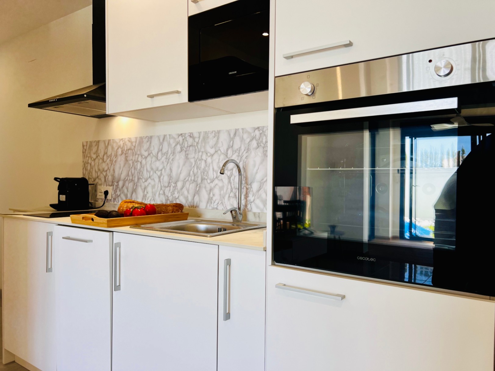 Moderne keuken met witte kasten, geïntegreerde Cecotec-apparatuur en een achterwand met marmereffect. Voorzien van een roestvrijstalen spoelbak en zwarte afzuigkap.
