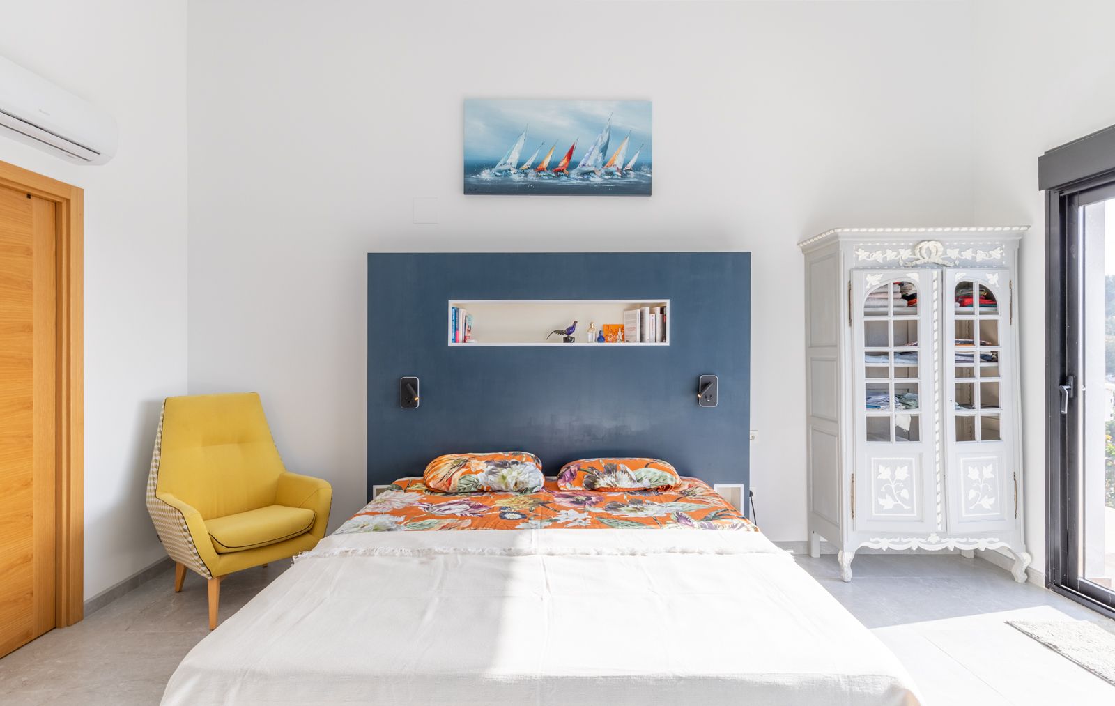 Moderne slaapkamer met een blauw hoofdbord met nis, een gele fauteuil en een klassieke kast. Voorzien van airconditioning en toegang tot het terras via een schuifpui.