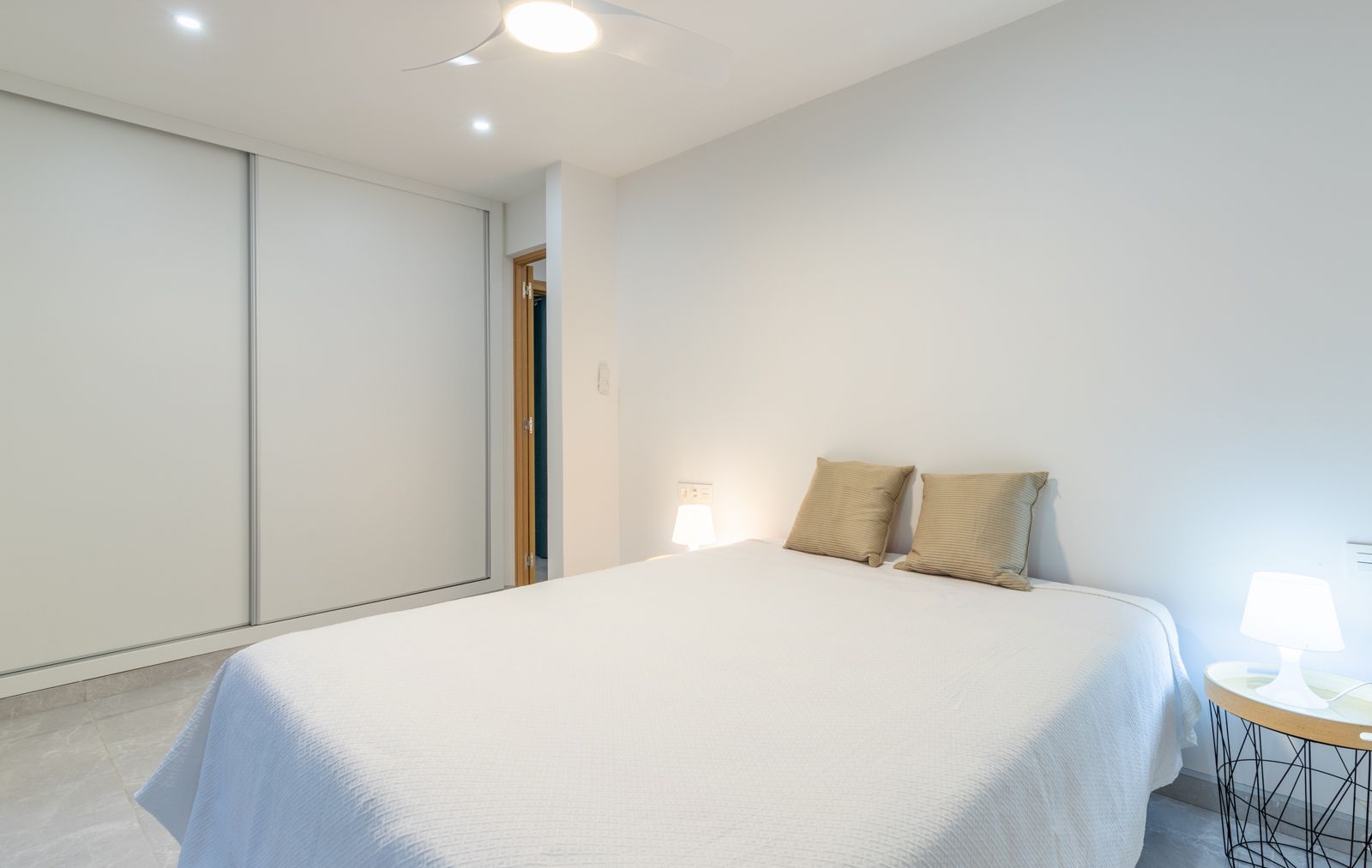 Moderne slaapkamer met een grote witte schuifdeurkast, grijze porseleinen tegelvloer en geïntegreerde plafondventilator in een minimalistische stijl.