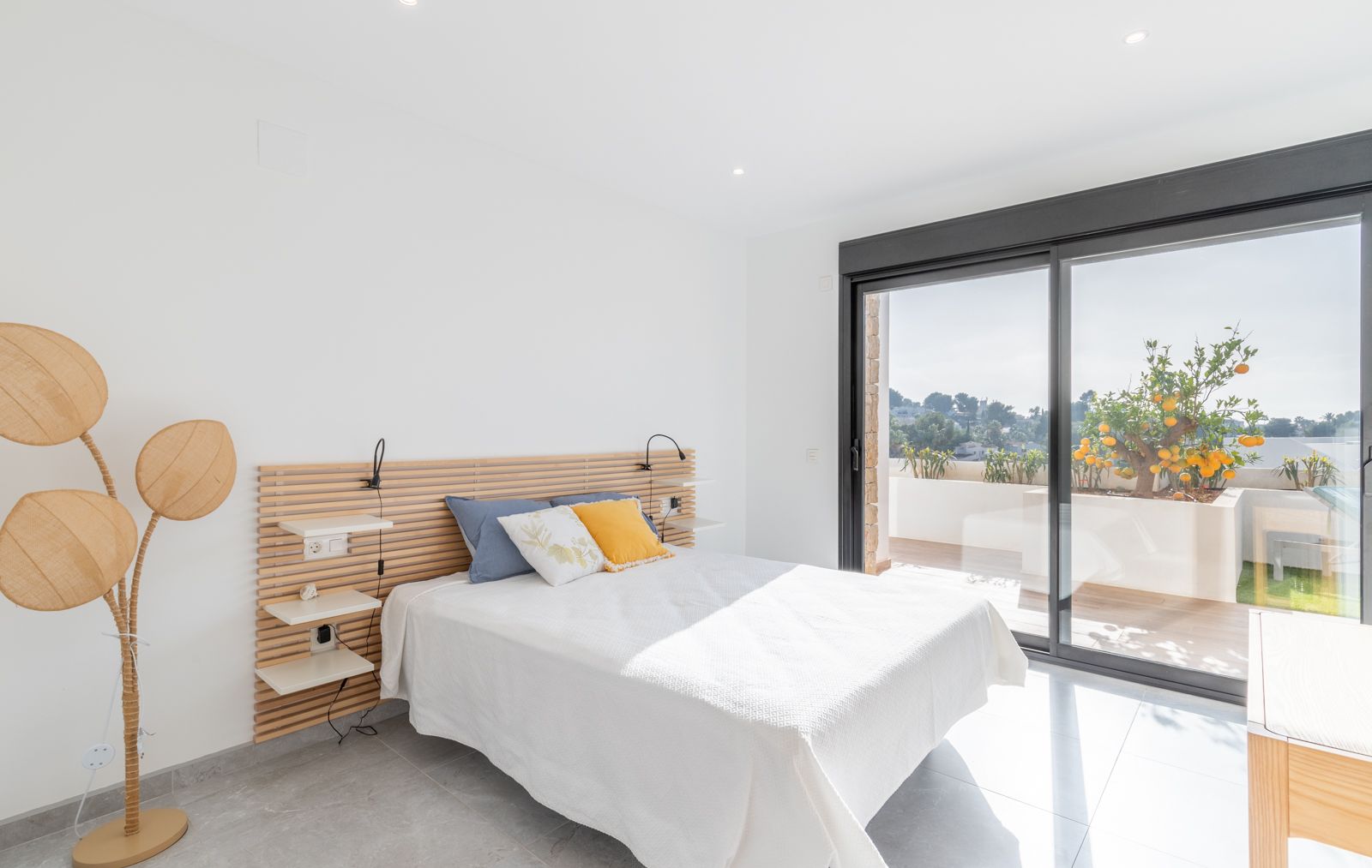 Moderne slaapkamer met een houten latten hoofdbord, geïntegreerde planken en grote schuifdeuren naar een terras met een citrusboom.