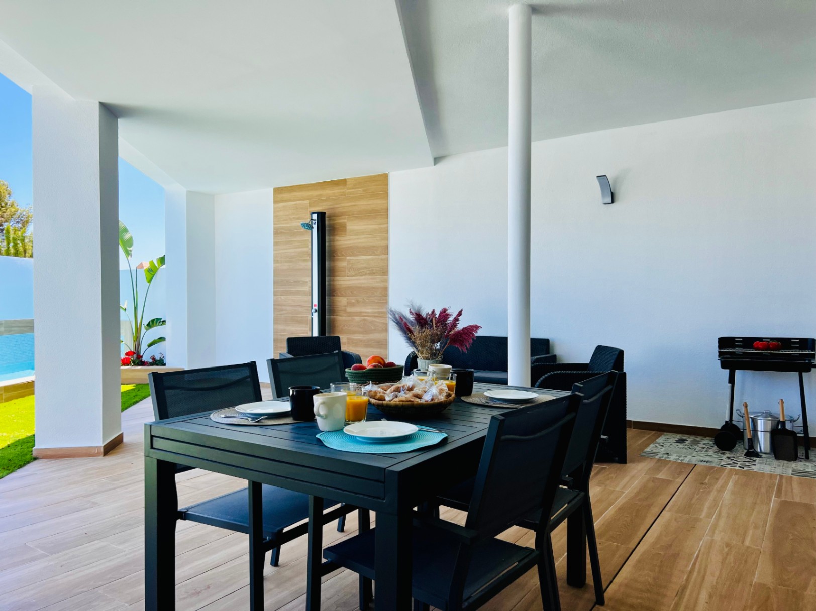 Overdekt terras met houtlook porseleinen vloer, een moderne eethoek, barbecueplek en een buitendouche geïntegreerd in een decoratief paneel.