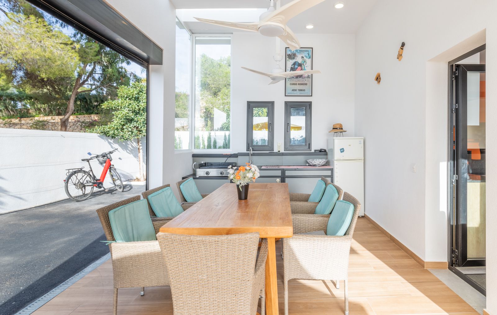 Overdekte buitenruimte met zomerkeuken, massief houten tafel, rotan stoelen en plafondventilatoren, direct verbonden met de zonnige buitenplaats.