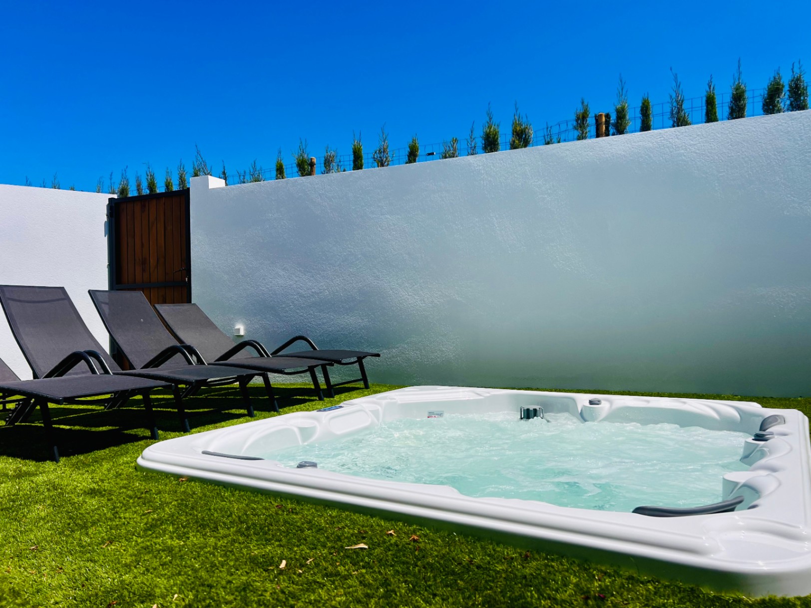 Privé-jacuzzi ingebouwd in kunstgras, voorzien van moderne ligbedden en een witte omheiningsmuur voor optimale privacy in de buitenlucht.