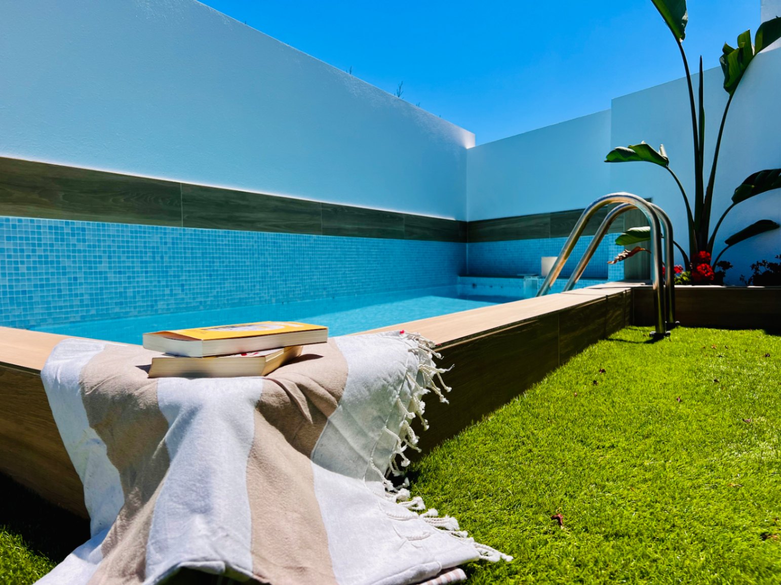 Privé jacuzzi met blauwe mozaïektegels, houtlook afwerking en kunstgrasveld, omgeven door strakke witte muren en een heldere blauwe lucht.
