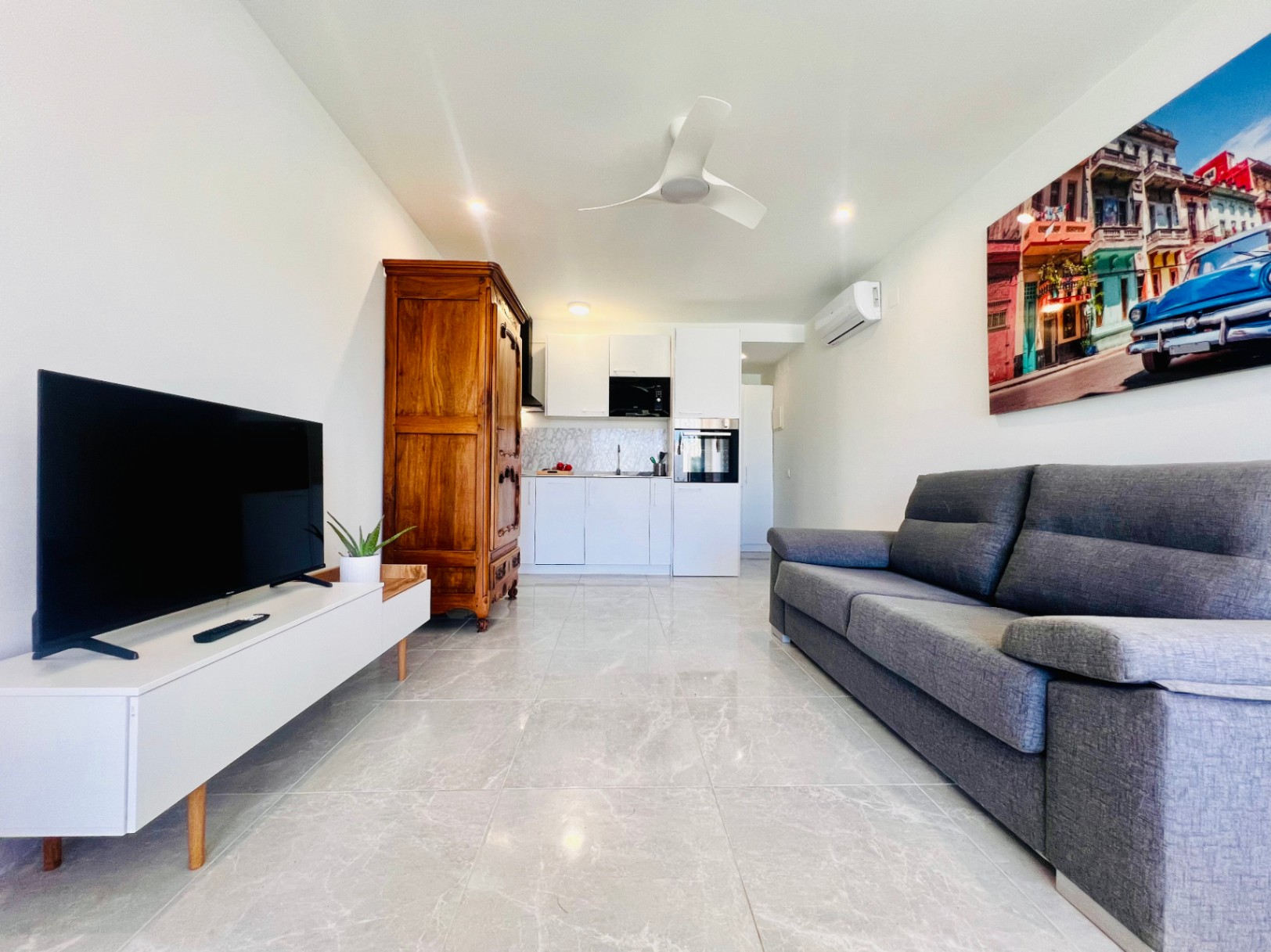 Woonkamer van een gastenverblijf met marmerlook vloertegels, een moderne witte kitchenette, grijze bank en een klassieke houten kast voor een uniek accent.