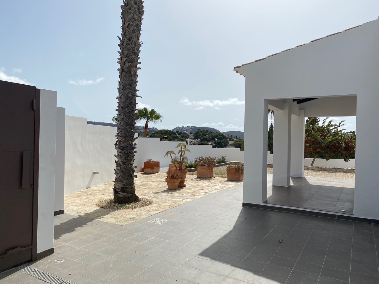 Buitenplaats met grijze vloertegels en natuursteen, voorzien van een palmboom en terracotta potten met uitzicht op de omliggende heuvels van Moraira.