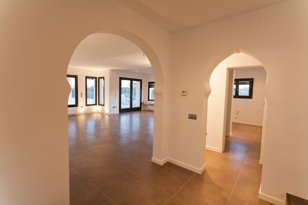 Hal met mediterrane bogen, terracotta-kleurige tegelvloer en doorkijk naar de woonkamer voorzien van donkere aluminium kozijnen.