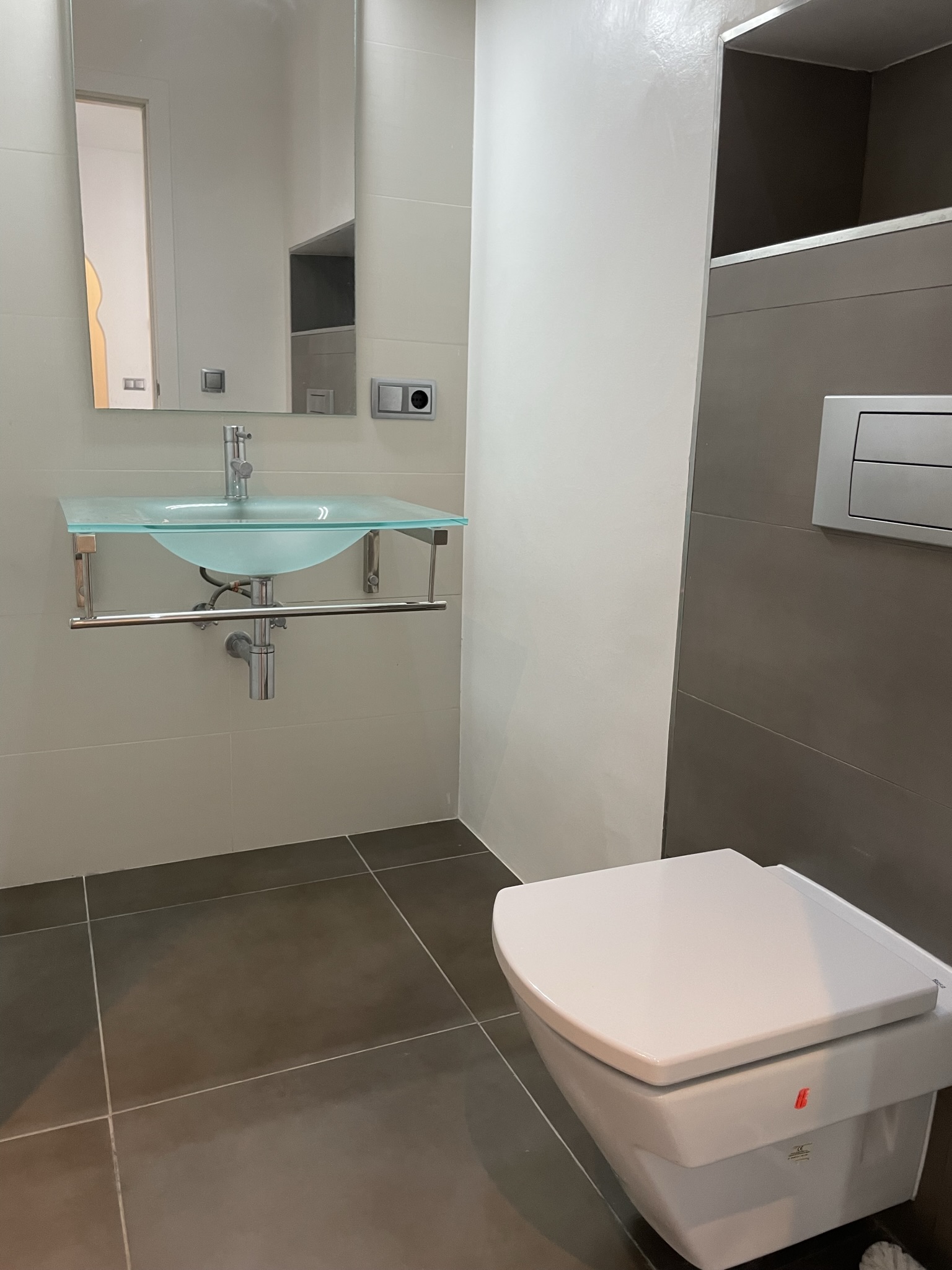 Moderne badkamer met een doorschijnende glazen wastafel op chromen steunen en een hangend toilet. Grootformaat grijze en crème tegels bieden een strakke afwerking.