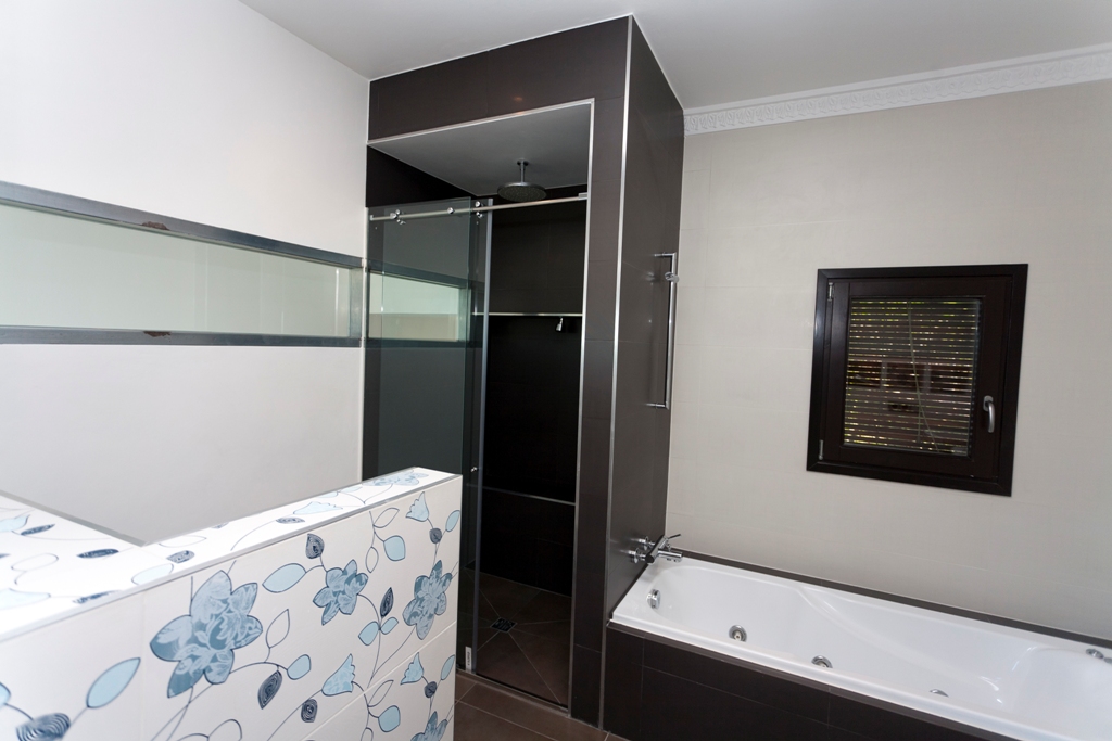 Moderne badkamer met ligbad en een aparte inloopdouche. Voorzien van chocoladebruine tegels, een decoratieve wand met bloemenprint en een raam met donker kozijn.