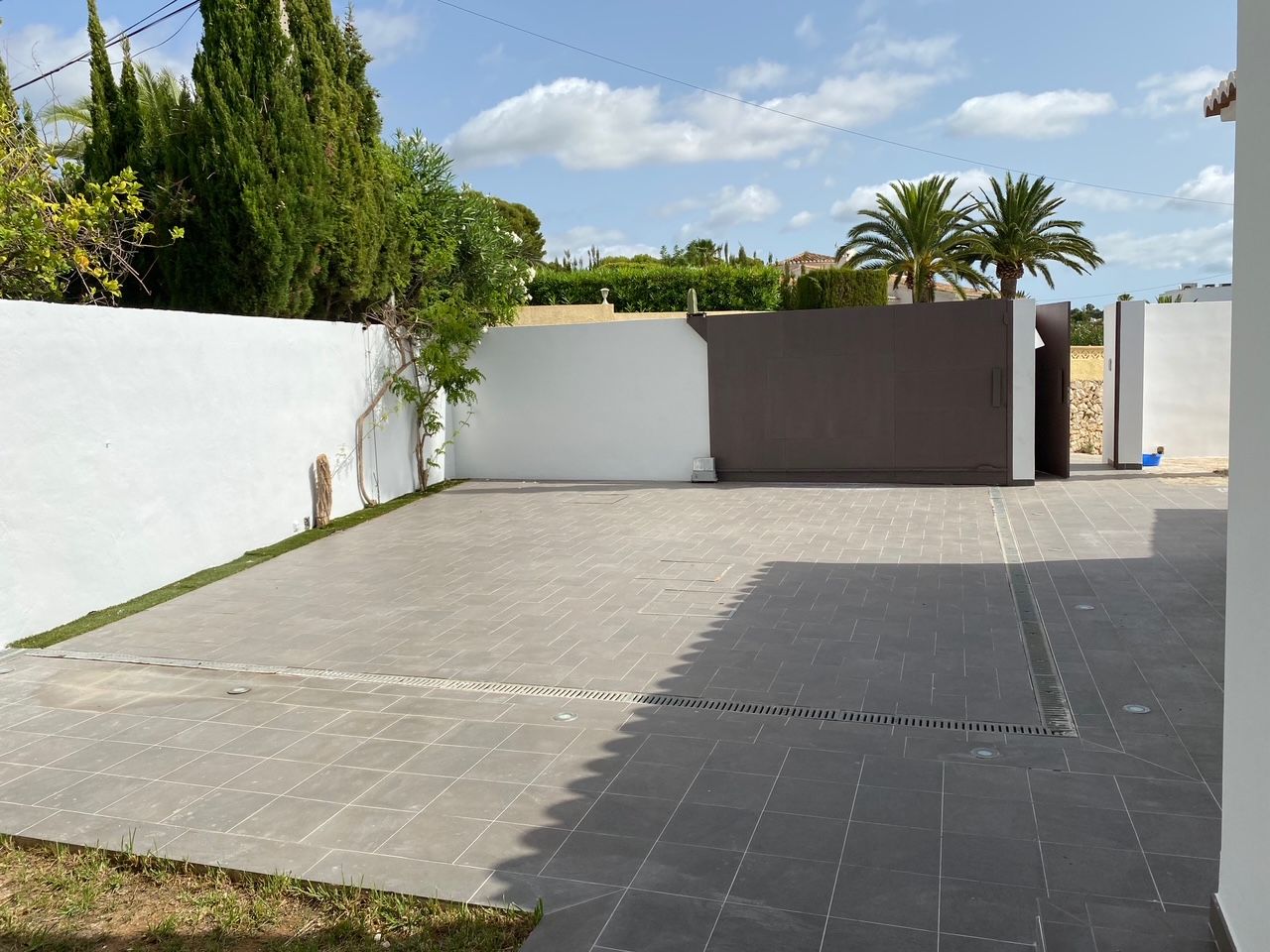Privé parkeerplaats met grijze tegels, een gemotoriseerde schuifpoort en aparte voetgangerstoegang, omsloten door witte muren en mediterrane beplanting.