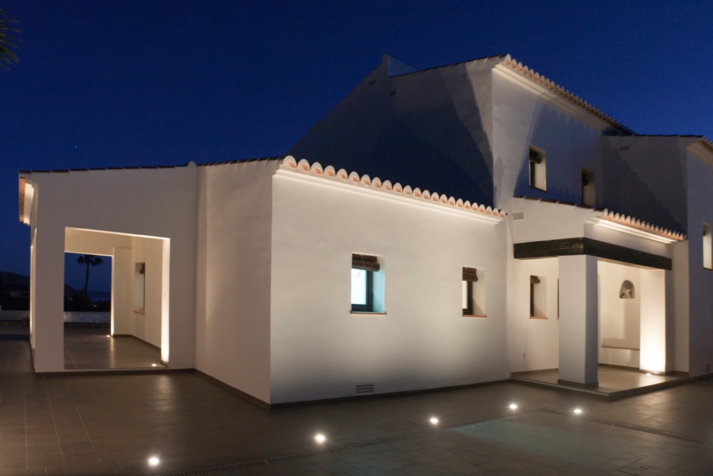 Witte mediterrane gevel verlicht bij schemering door vloerspots. Voorzien van traditionele dakpannen en een overdekte veranda met donkere houten balken op een betegeld terras.