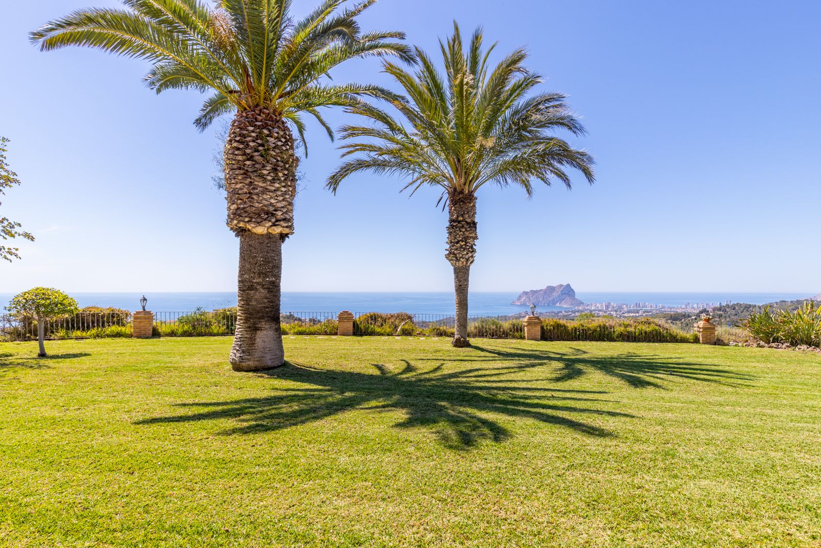 Uitzicht op de tuin met groen gazon, palmbomen en uitzicht op de Middellandse Zee en de Peñón de Ifach.