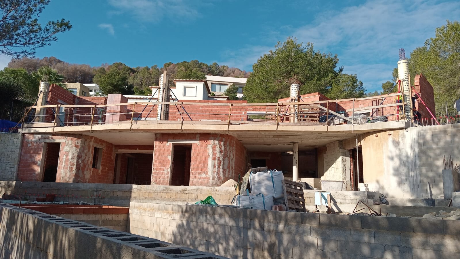 Gezicht op de ruwbouw van de villa, met het gebogen terras en de bakstenen muren van de twee woonlagen op een hellend perceel.