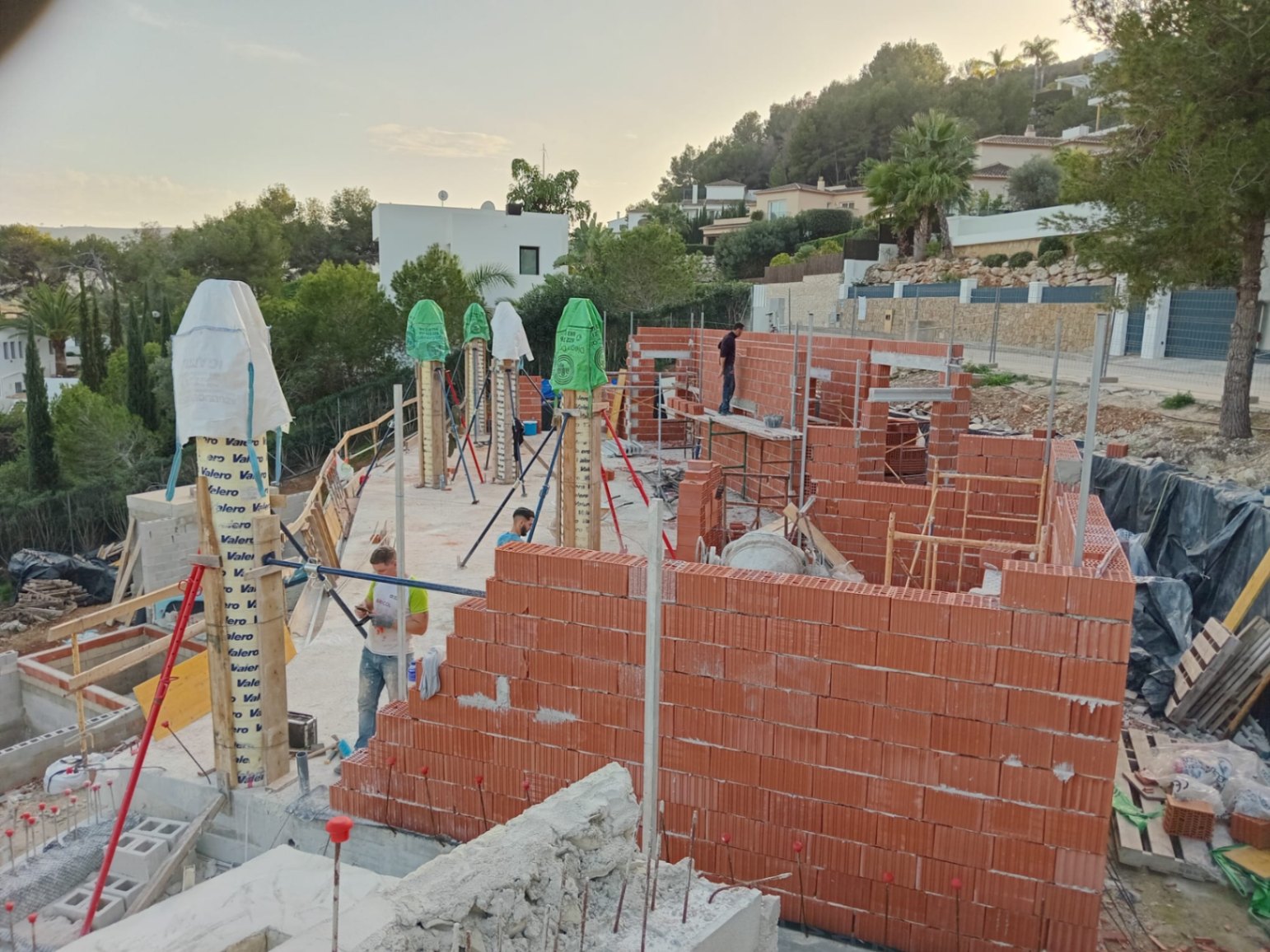 Huidige bouwfase met de constructie van bakstenen muren en structurele kolommen op een verhoogd perceel met uitzicht op de groene heuvels van Jávea.