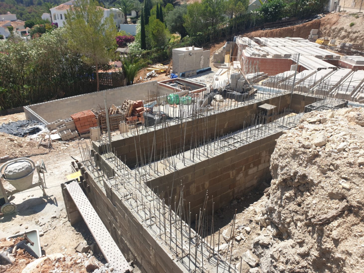 Vroege bouwfase met funderingsmuren, staalwapening en betonblokken op een verhoogd perceel omgeven door een natuurlijke mediterrane omgeving.