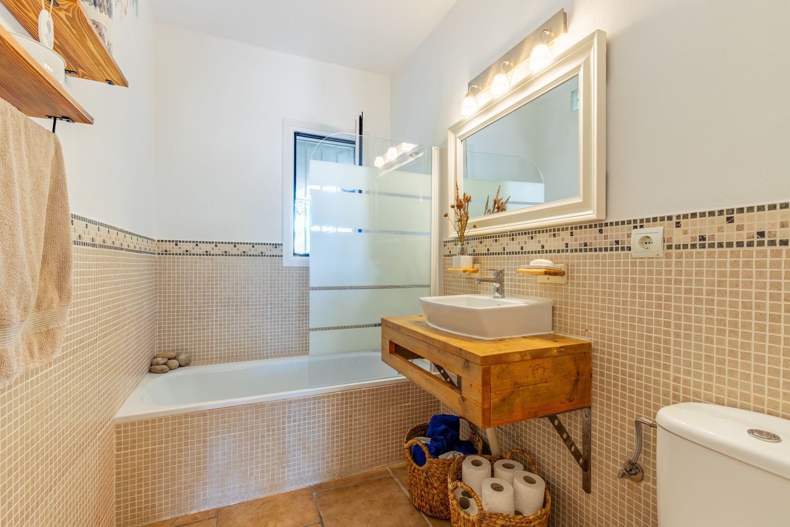 Badkamer in rustieke stijl met een massief houten wastafelmeubel, opzetwastafel, ligbad met glazen douchescherm en beige mozaïekwandtegels.