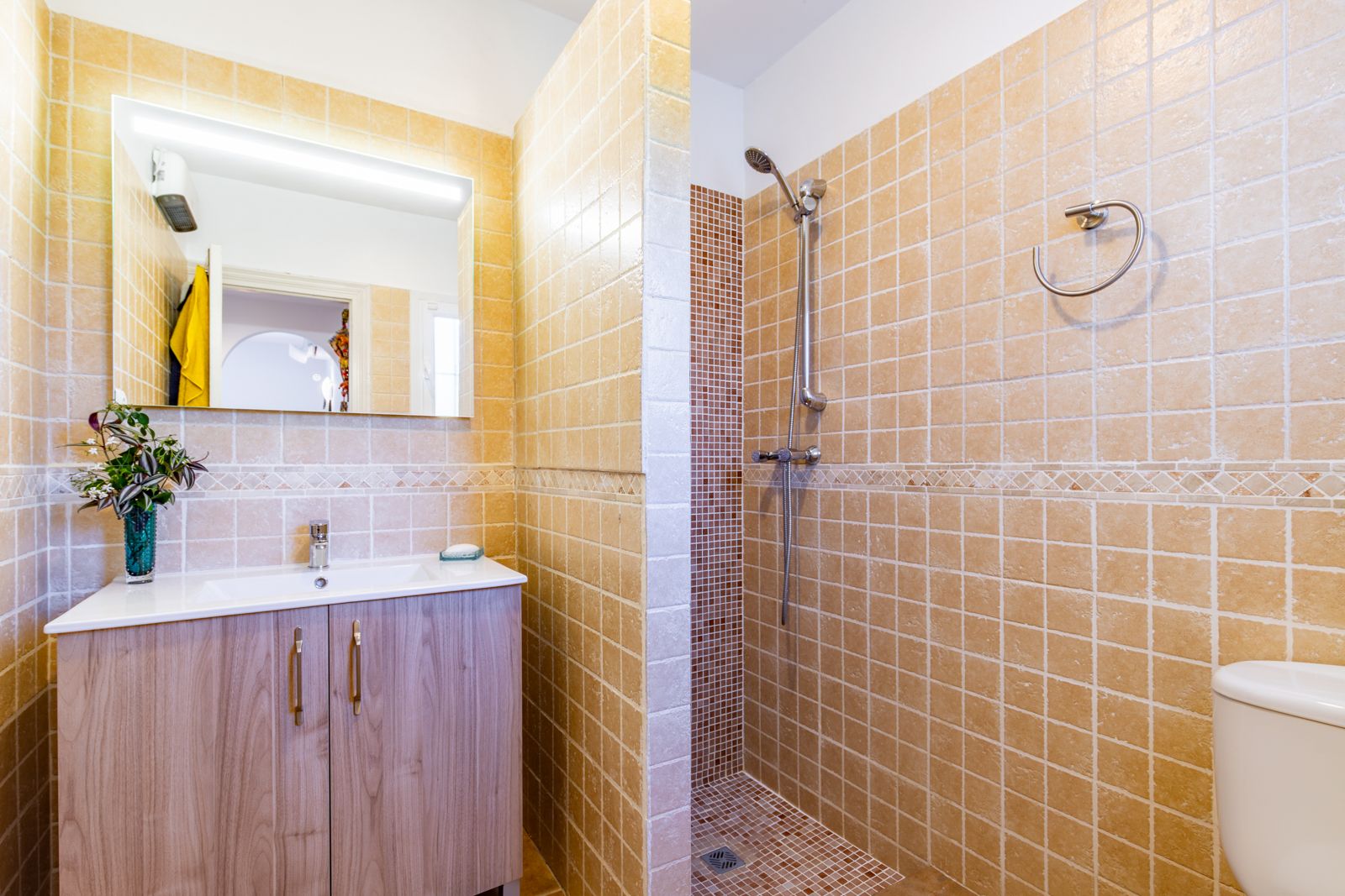 Badkamer met inloopdouche voorzien van mozaïekvloer en beige tegels. Inclusief een wastafelmeubel met houtnerf-afwerking en een spiegel met verlichting.