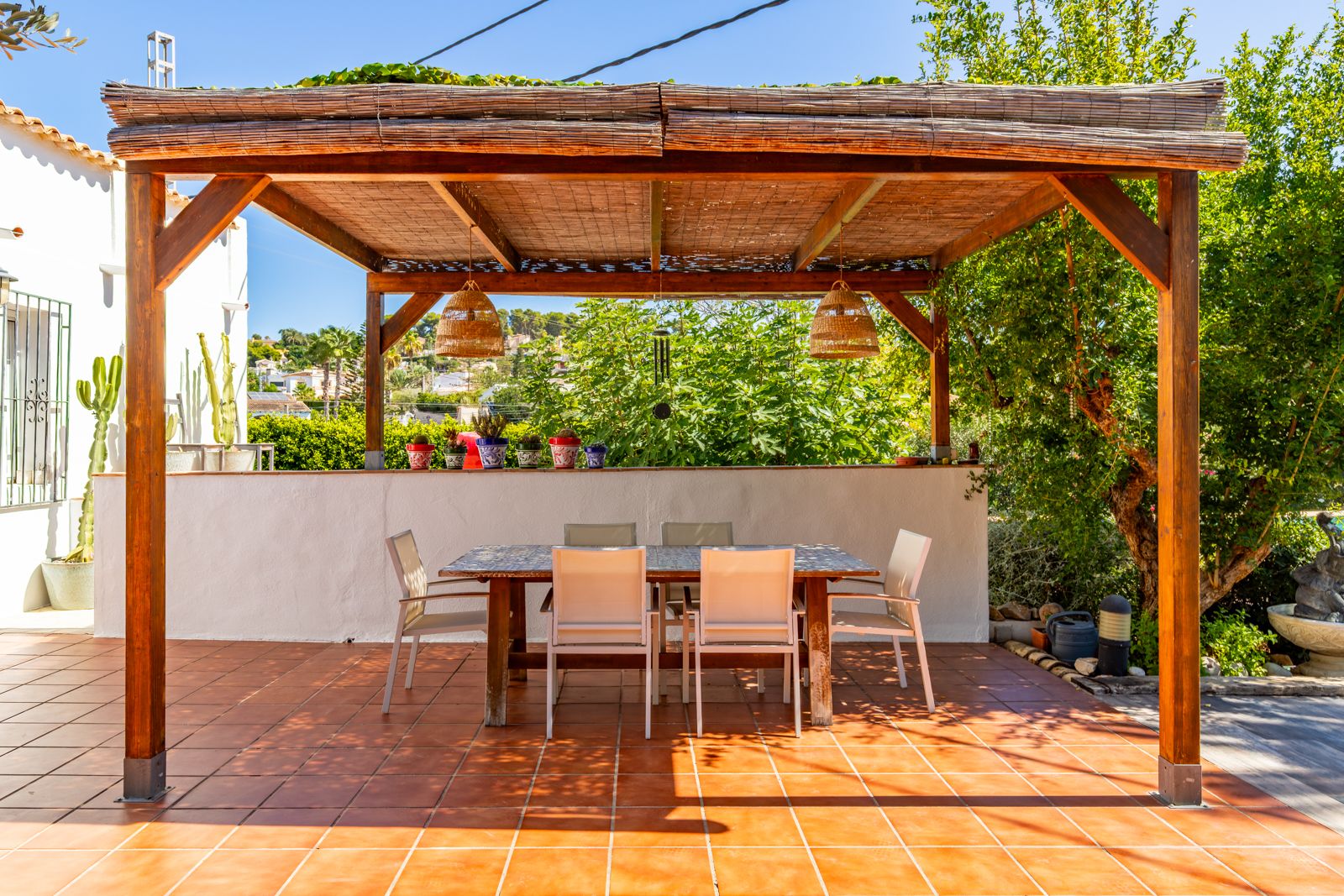 Buiten-eethoek onder een houten pergola met rieten dakbedekking, rotan hanglampen en een betegelde tafel op een terracotta terras.