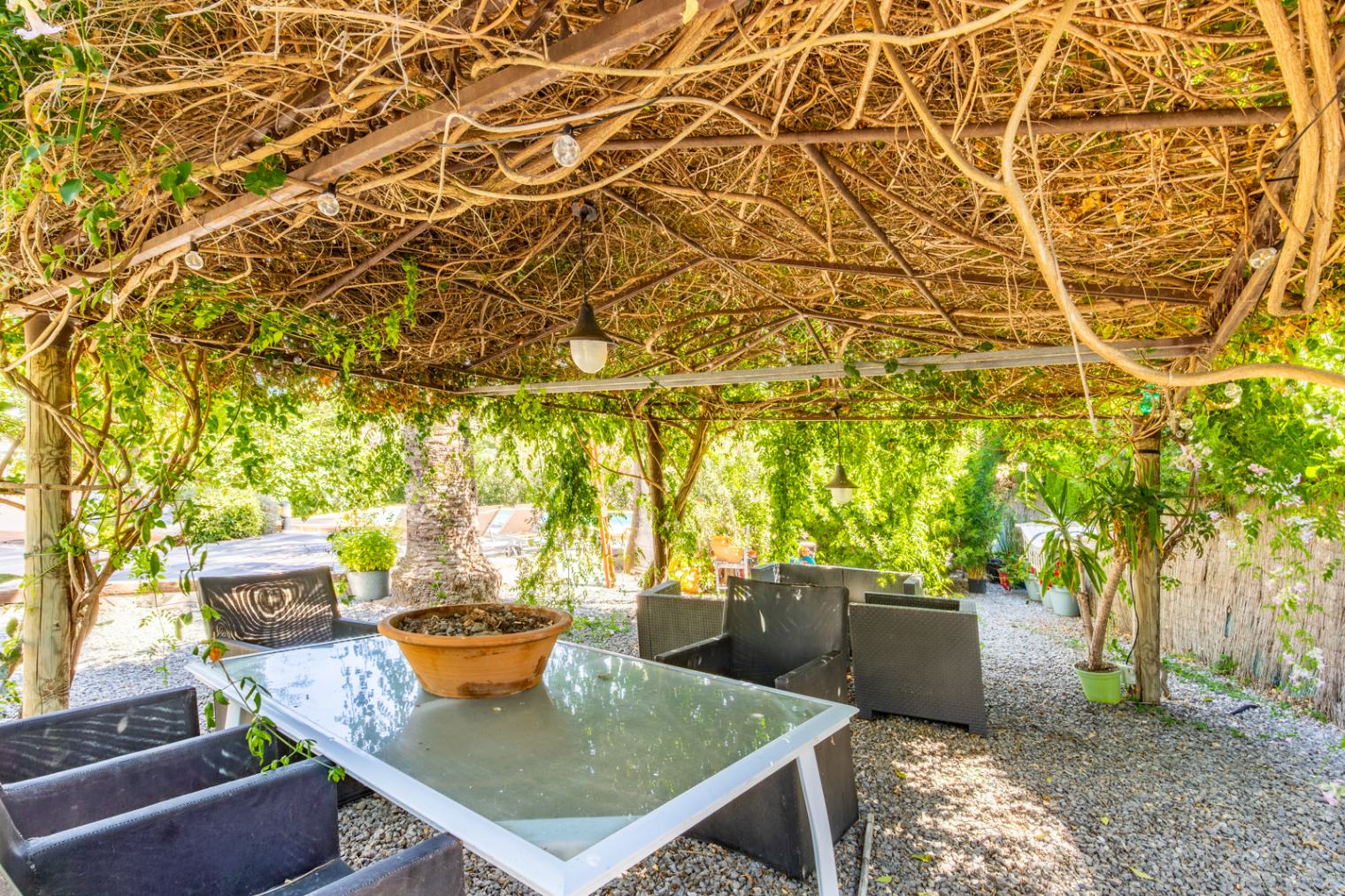 Buiten-eethoek onder een natuurlijke pergola met klimplanten, voorzien van een glazen tafel, rotan stoelen en een grindvloer in een volgroeide mediterrane tuin.