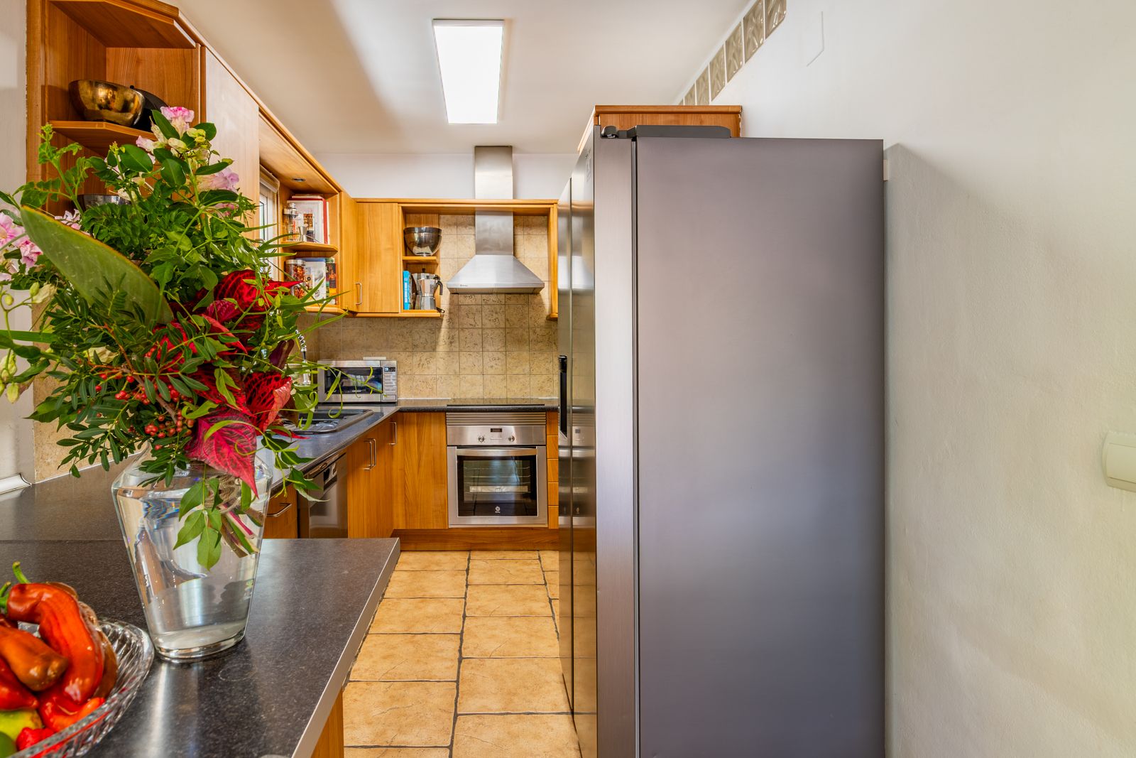 Functionele keuken met houten kasten, donker werkblad en roestvrijstalen apparatuur. Voorzien van een terracotta tegelvloer en een grote Amerikaanse koelkast.