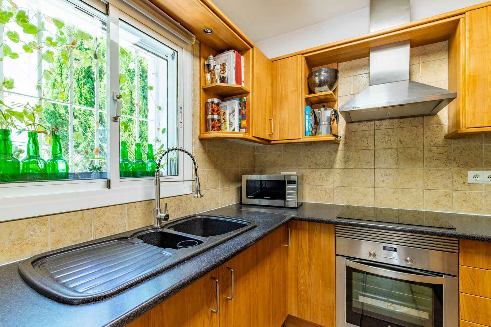 Ingerichte keuken met houten kasten, een donker granieten werkblad en dubbele spoelbak. Voorzien van rvs-apparatuur en natuurlijk licht via het tuinraam.