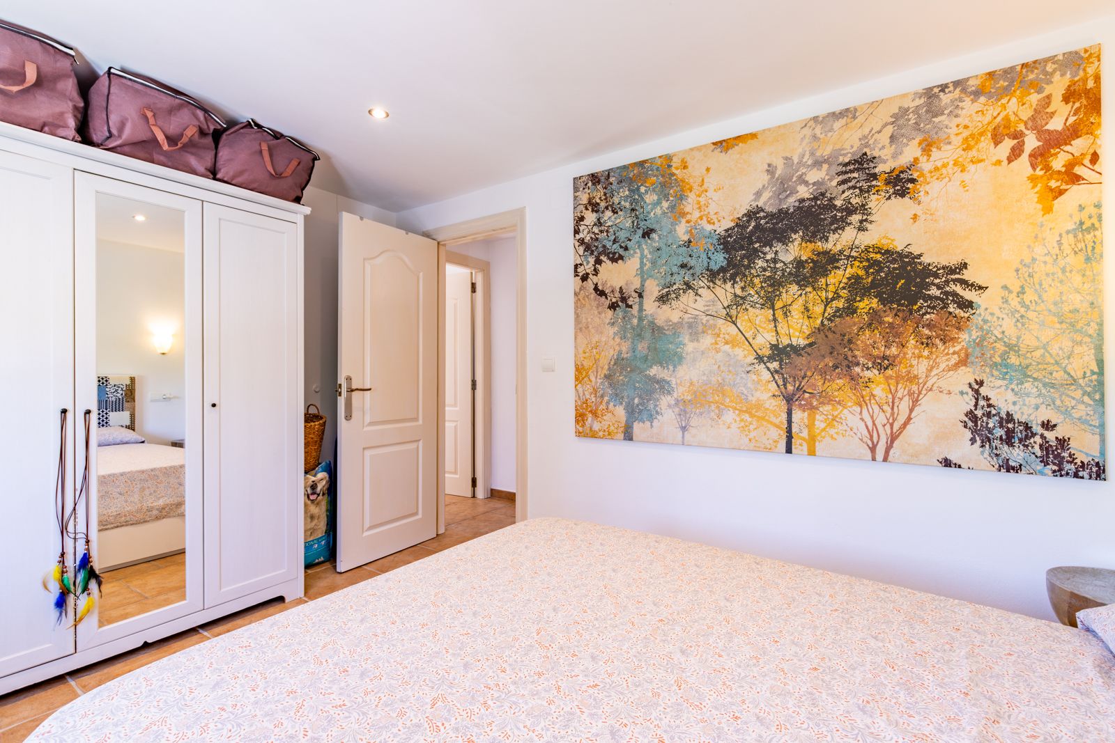 Lichte slaapkamer met een witte driedeurs kledingkast met spiegel en een groot decoratief wandpaneel met natuurmotief. Voorzien van tegelvloer en inbouwspots.