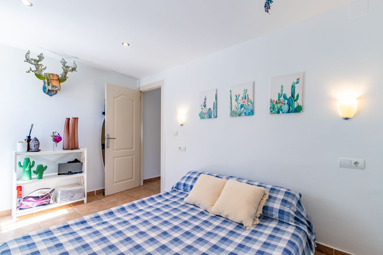 Lichte slaapkamer met terracotta vloertegels, een bed met blauw geruit textiel en cactusdecoratie. Voorzien van inbouwspots en warme wandarmaturen.
