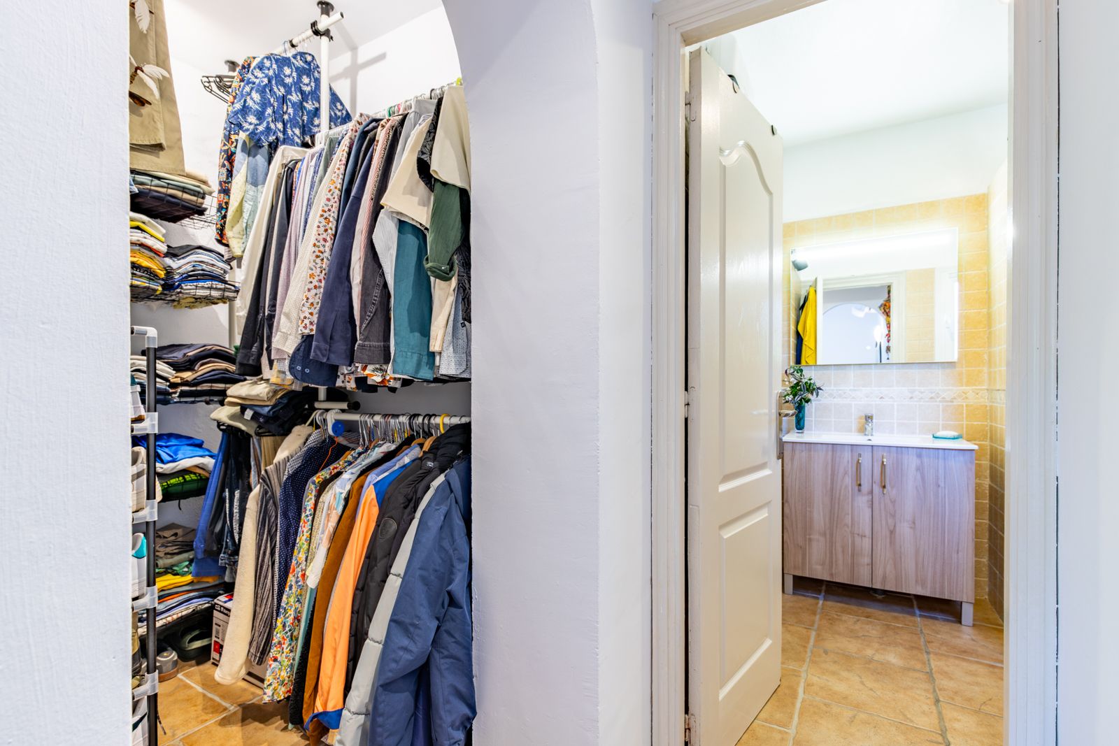 Open inloopkast met legplanken en hangrails, grenzend aan een badkamer voorzien van een houten wastafelmeubel en verlichte spiegel.