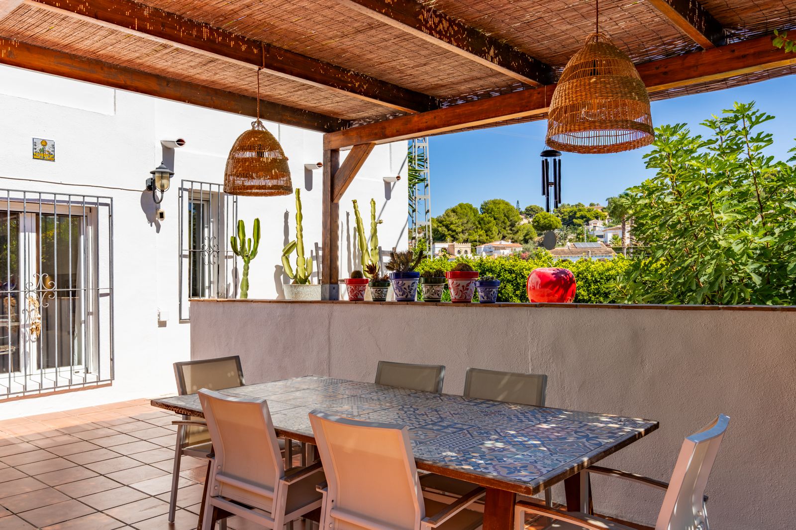 Overdekt terras met houten pergola en rieten dak, voorzien van een betegelde eettafel en rieten hanglampen met uitzicht op de groene omgeving.