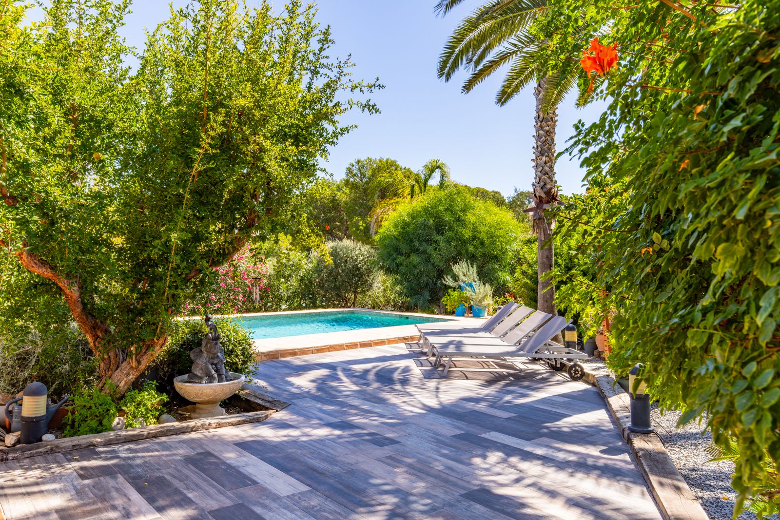 Zonnig terras met houtlook tegels, grijze ligbedden en een privézwembad, omgeven door volgroeide mediterrane beplanting en palmbomen.