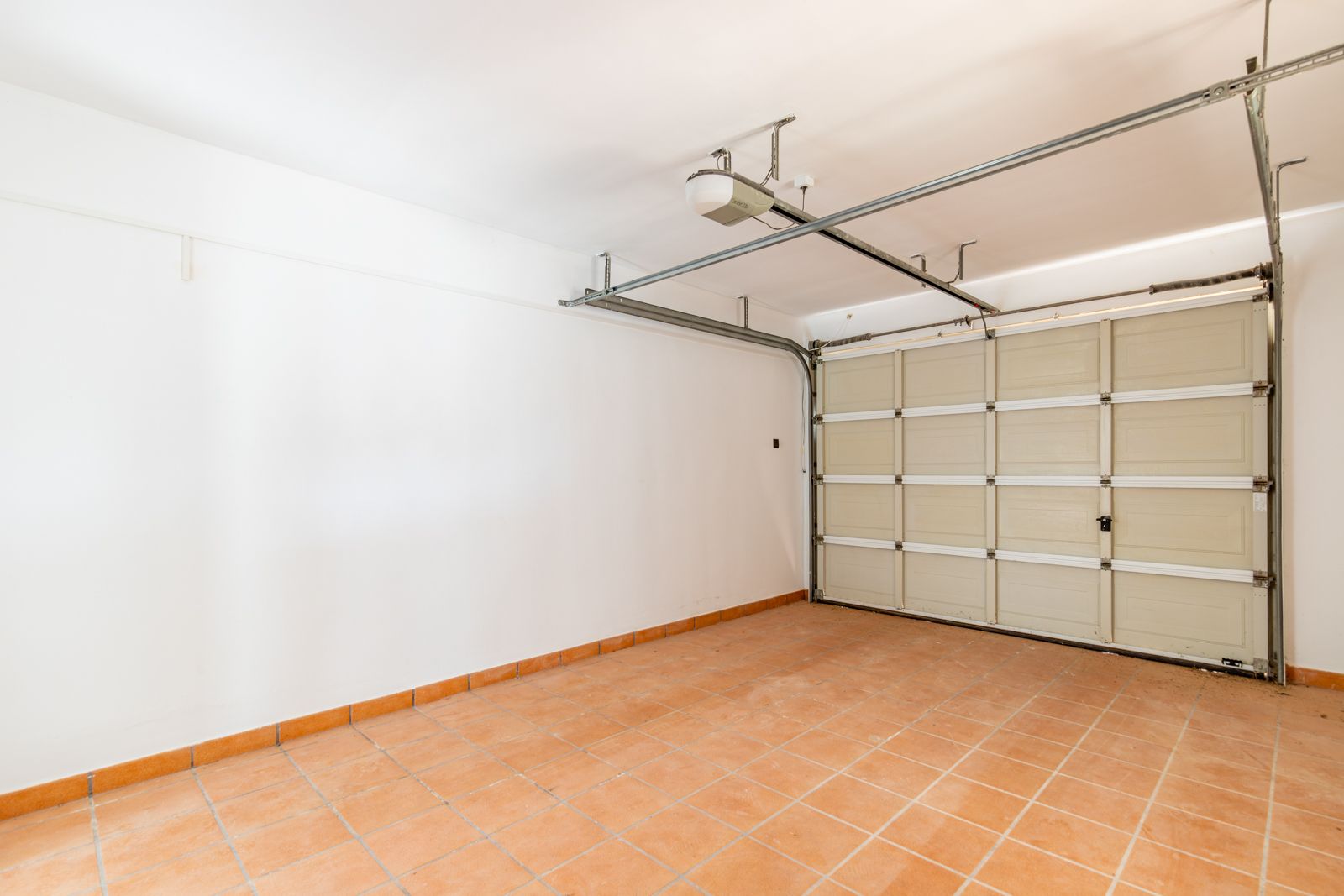 Afgesloten garage met terracotta-stijl keramische vloertegels, gladde witte muren en een gemotoriseerde sectionaaldeur met een automatisch openingssysteem aan het plafond.