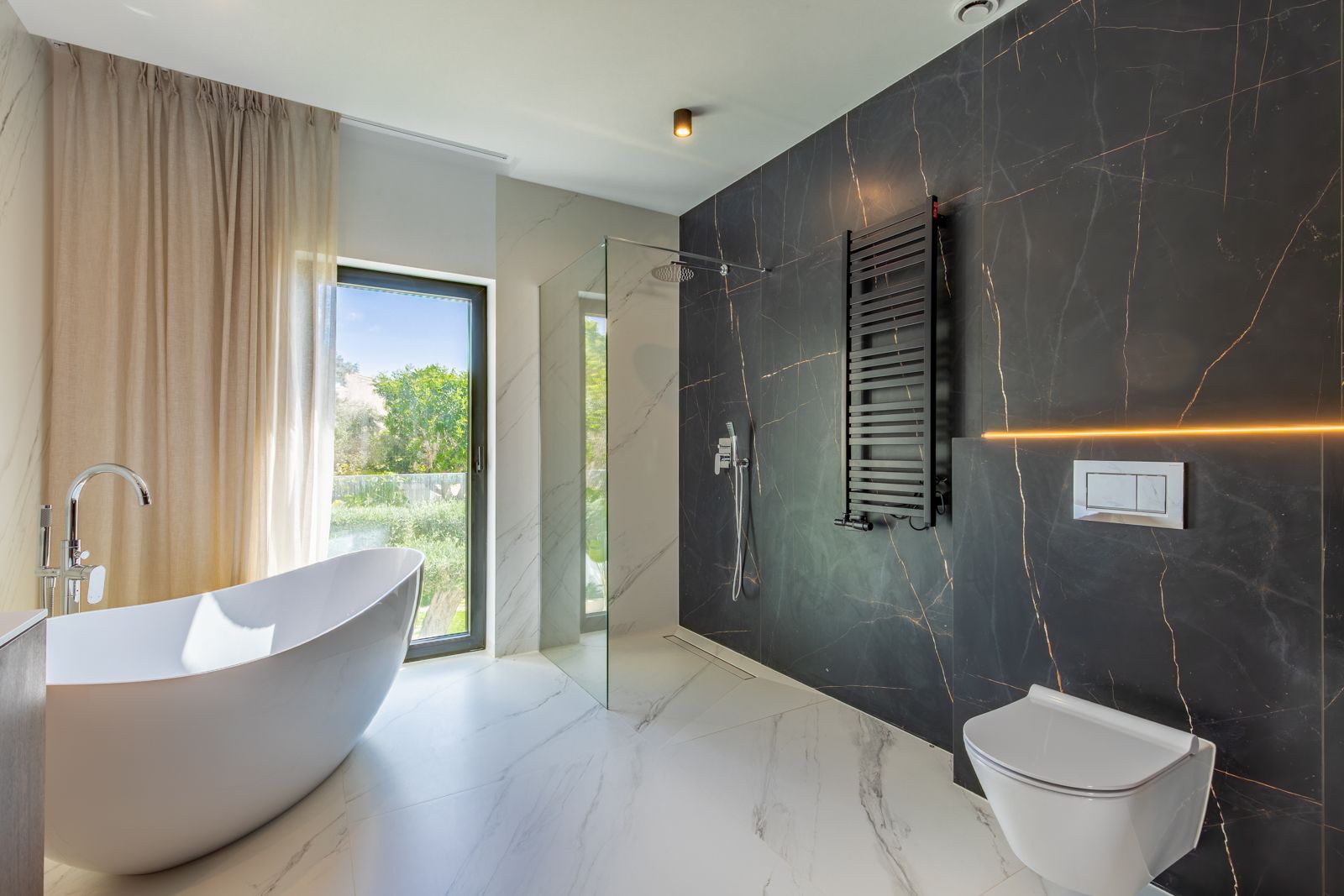 Designbadkamer met vrijstaand ligbad, inloopdouche en contrasterende porseleinen wandpanelen. Voorzien van een hangend toilet, zwarte handdoekradiator en uitzicht op de tuin.