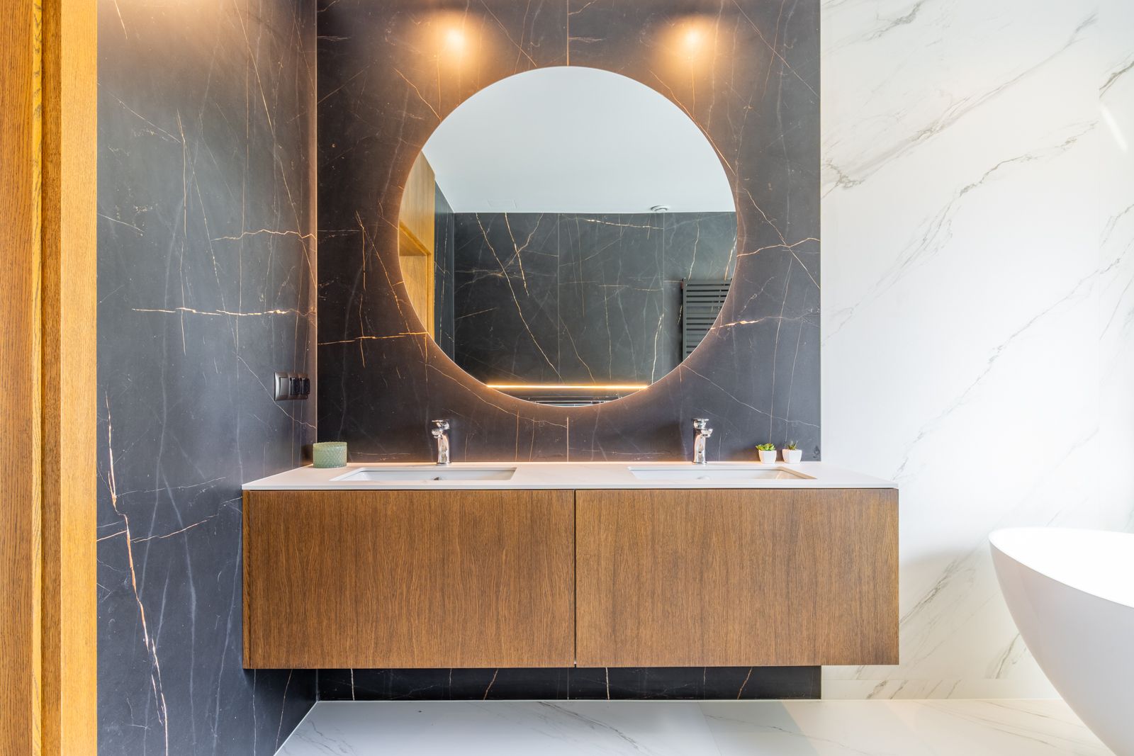 Designbadkamer met zwevend eikenhouten meubel, dubbele wastafel en ronde achterverlichte spiegel tegen een wand van donker porselein met gouden aders.