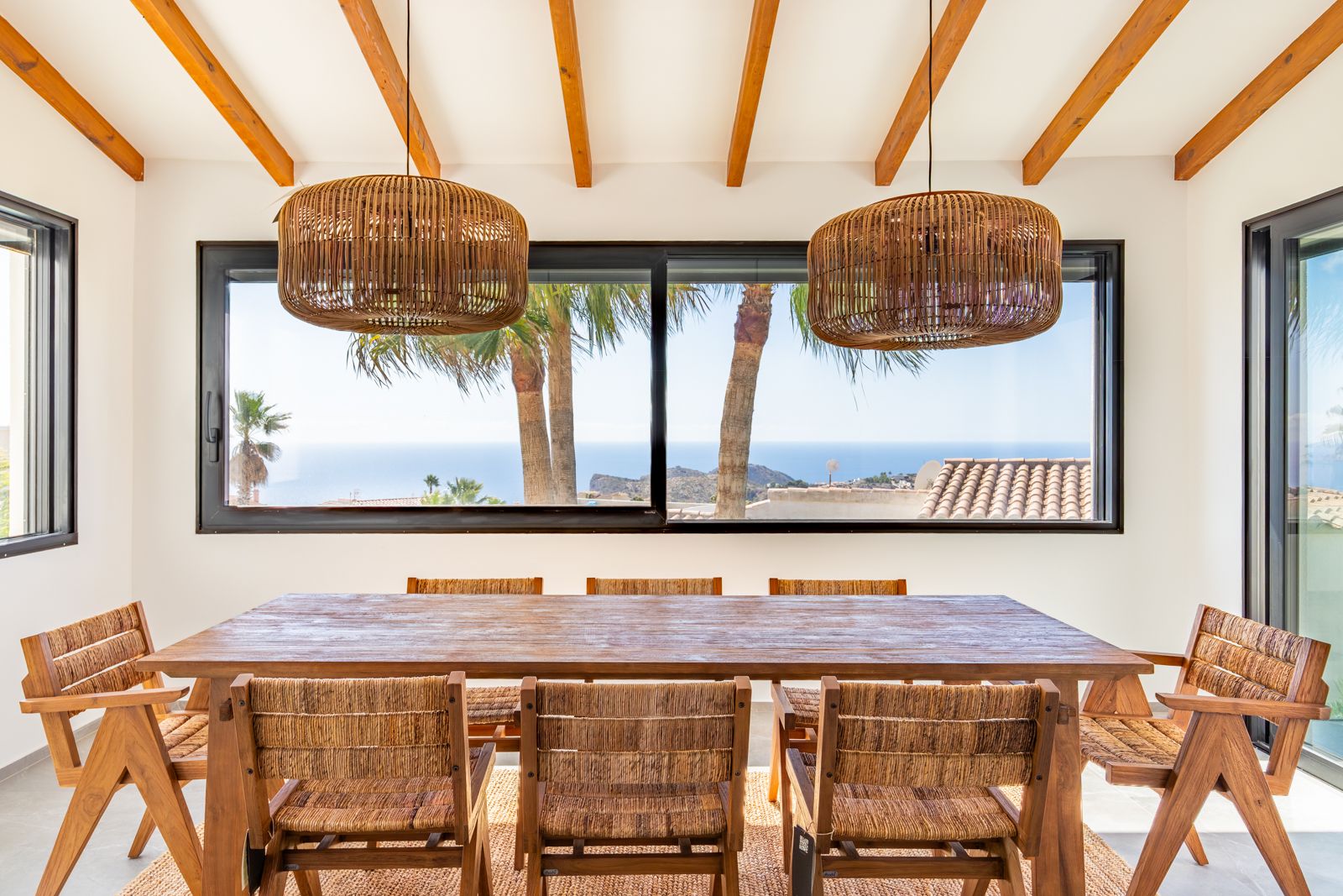 Eetkamer met houten balkenplafond, massieve tafel en rotan lampen. Het grote raam biedt een weids uitzicht op de Middellandse Zee en omliggende palmen.