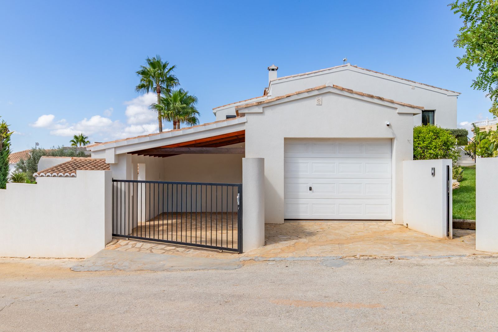 Hoofdingang met een gesloten garage en een overdekte carport. Voorzien van natuurstenen bestrating, een zwart metalen hek en witte mediterrane architectuur.
