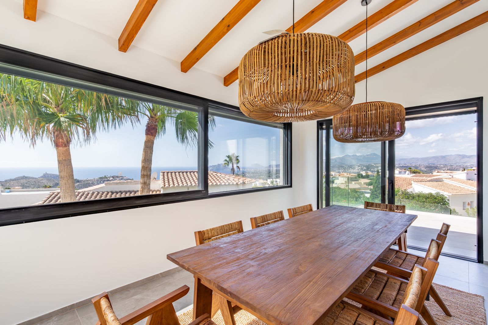 Lichtrijke eetkamer met houten balkenplafond, massief houten tafel en rotan hanglampen. Grote raampartijen bieden panoramisch uitzicht op de Middellandse Zee en de bergen.