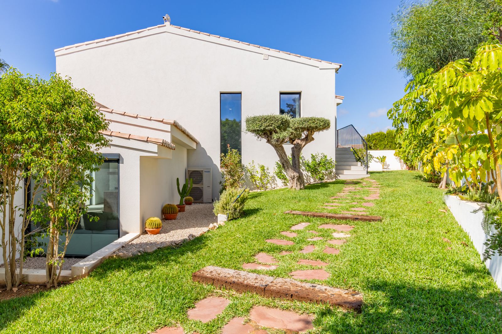 Mediterrane tuin met verzorgd gazon, een karakteristieke olijfboom en een pad van natuursteen langs de strakke witte gevel van de villa.