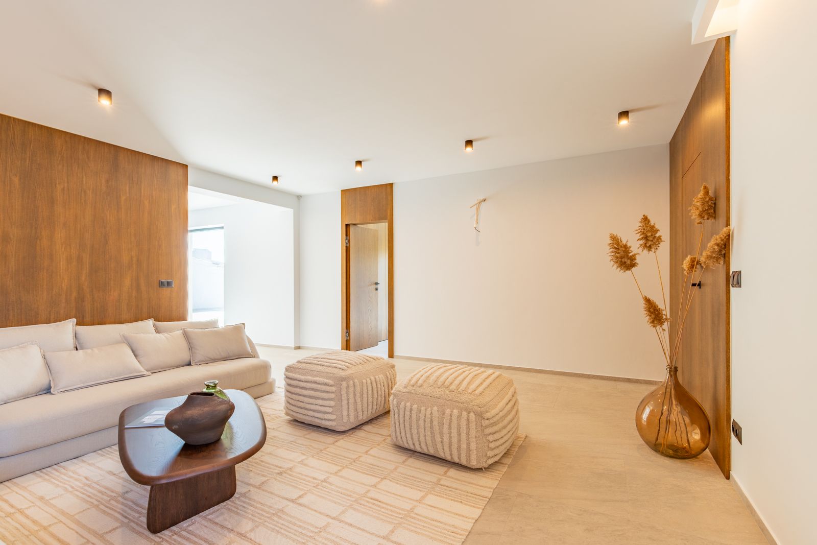 Minimalistische woonkamer met wandpanelen van natuurlijk eikenhout, grootformaat porseleinen vloeren en designmeubilair verlicht door subtiele inbouwspots.