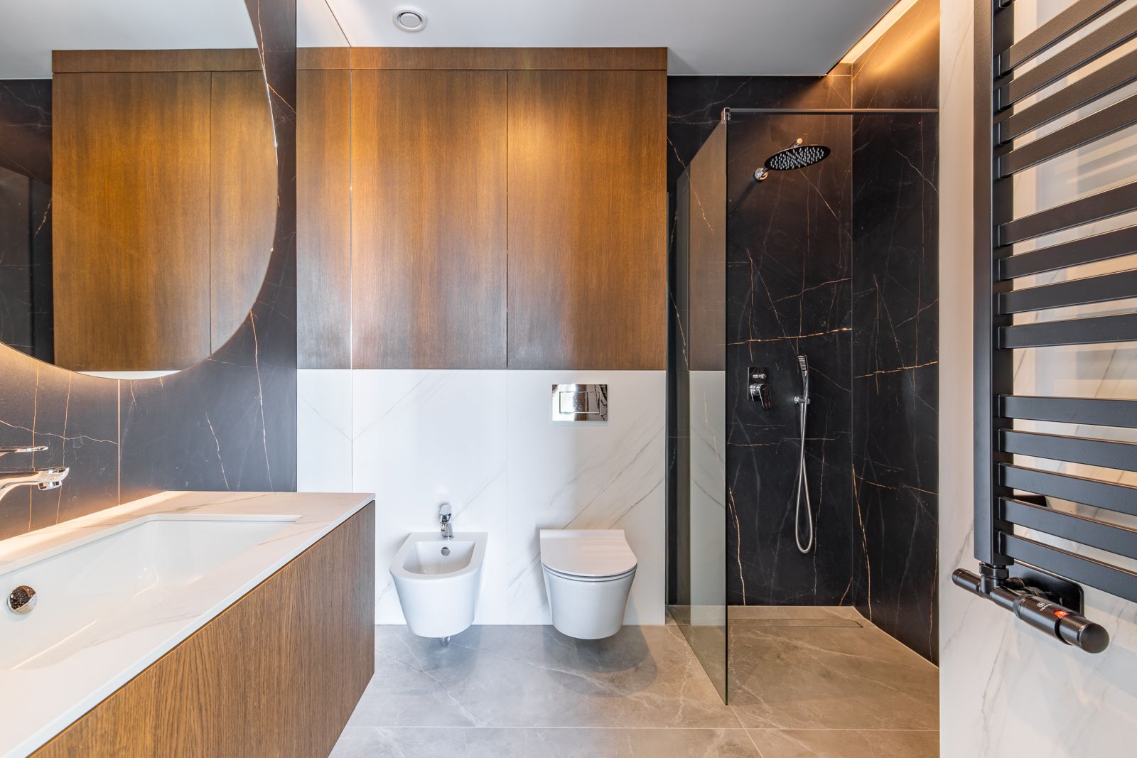 Moderne badkamer met donkere porseleinen tegels, inloopdouche, hangend sanitair en natuurlijk eikenhouten meubilair onder een grote verlichte ronde spiegel.