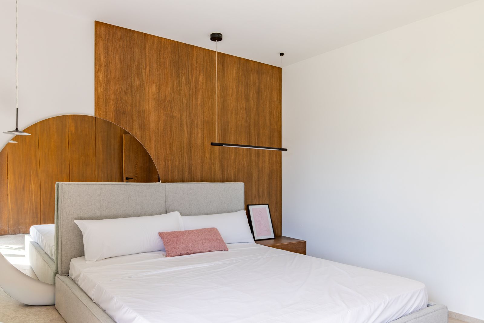 Moderne slaapkamer met wandpanelen van natuurlijk eikenhout, een grijs stoffen hoofdbord en een grote ronde spiegel. Minimalistische zwarte hanglampen en geïntegreerd nachtkastje.