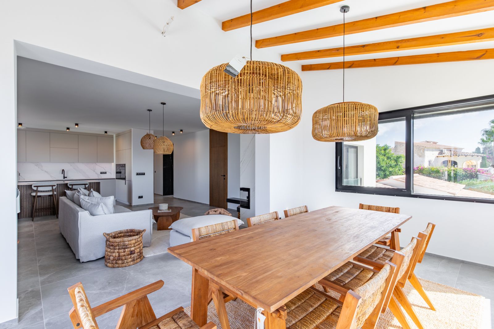 Open eetkamer met massief houten tafel, zichtbare plafondbalken en rotan hanglampen, verbonden met de woonkamer en moderne keuken.