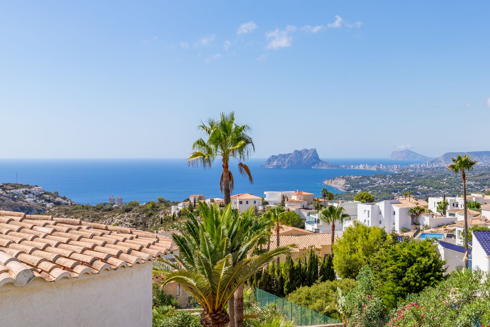 Panoramisch uitzicht op de Middellandse Zee en de Peñón de Ifach, met kustarchitectuur en weelderige palmbomen onder een heldere hemel.