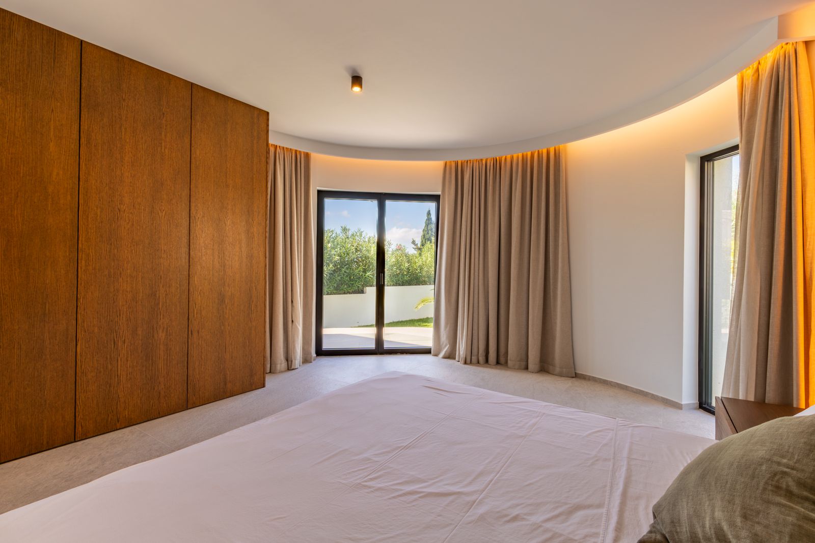 Slaapkamer met gebogen wand, op maat gemaakte eikenhouten kasten en indirecte verlichting. Grote schuifdeuren verbinden de ruimte direct met buiten.
