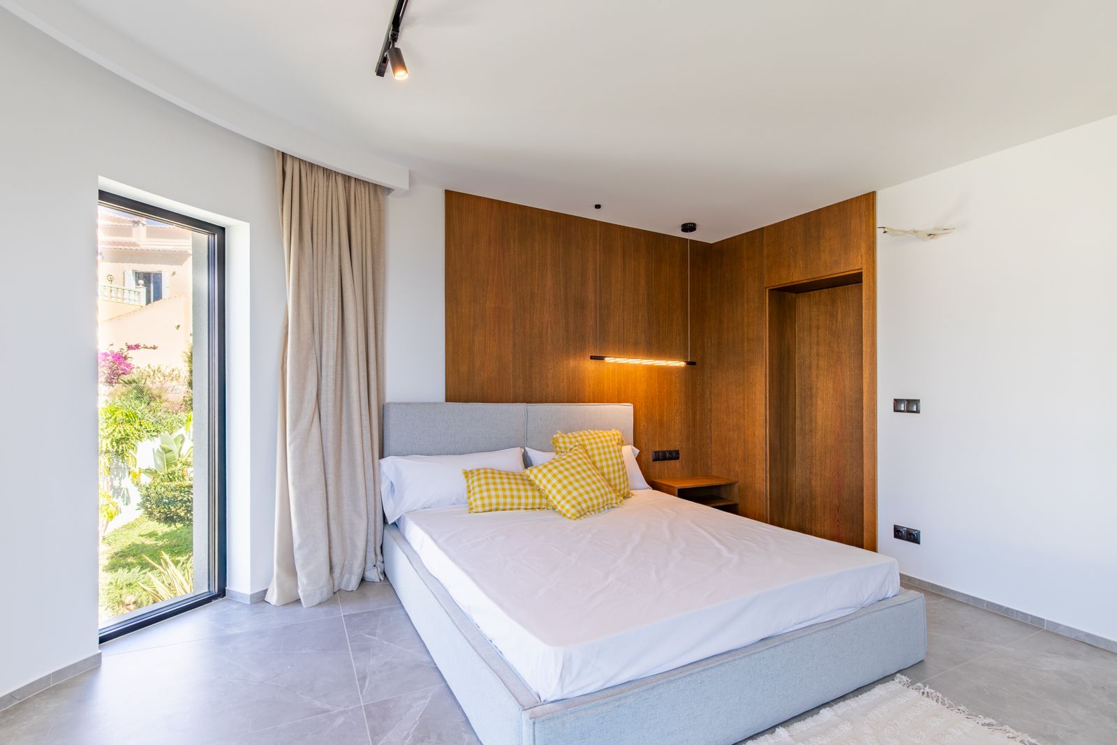 Slaapkamer met natuurlijke eikenhouten wandpanelen, design hangverlichting en grootformaat porseleinen vloertegels. De grote raampartij kijkt uit op de mediterrane tuin.