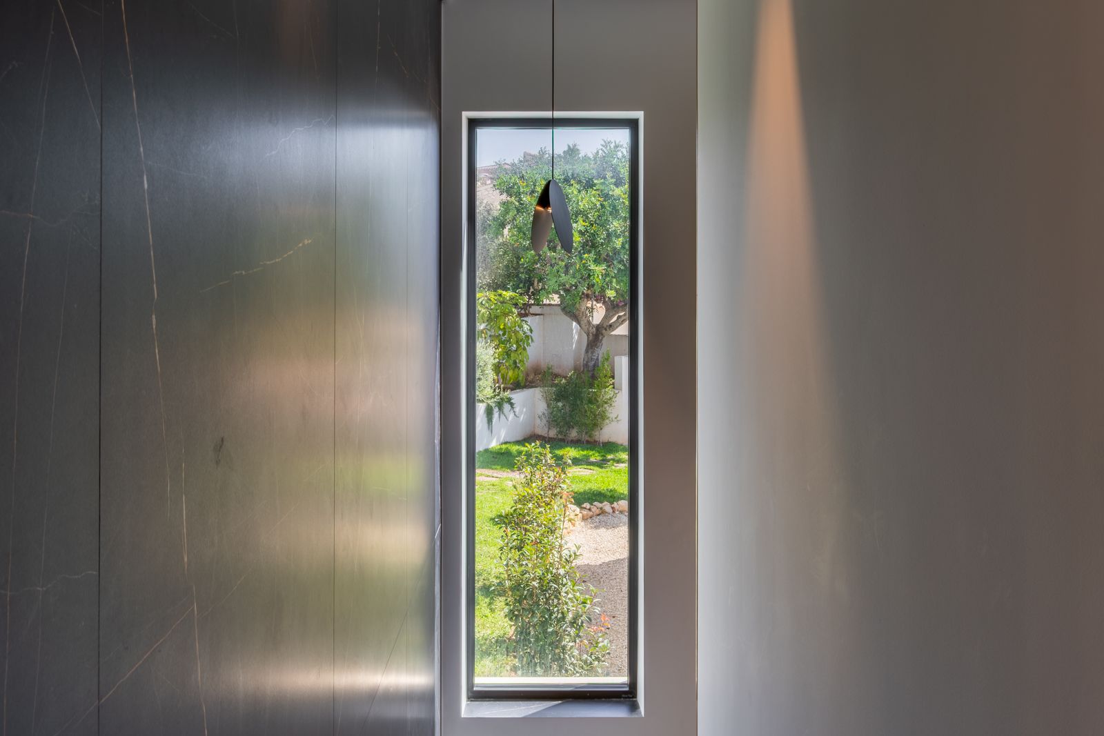 Verticaal raam met drievoudige beglazing en zicht op de tuin, gecombineerd met donkere grootformaat porseleinen wandpanelen en een minimalistische designlamp.