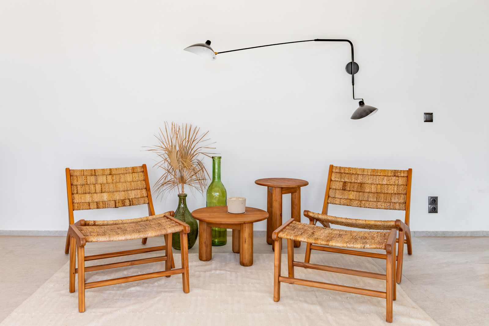 Zithoek met houten fauteuils en natuurlijk vlechtwerk, teakhouten tafels en een zwarte design wandlamp op een lichte keramische tegelvloer.