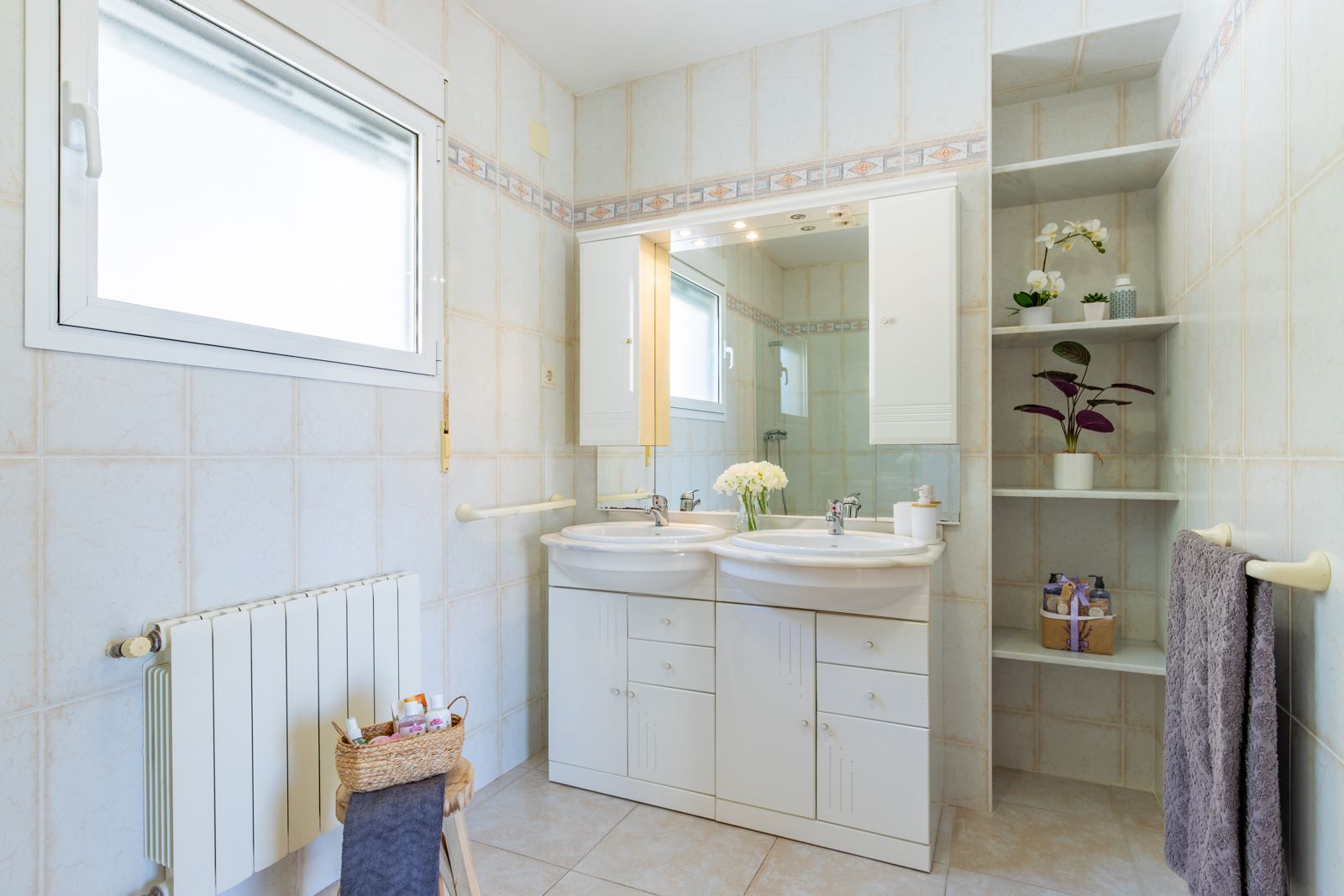 Badkamer met dubbele wastafel, wit badkamermeubel, verlichte spiegel en ingebouwde open schappen. Betegelde wanden met sierrand en een wandradiator.