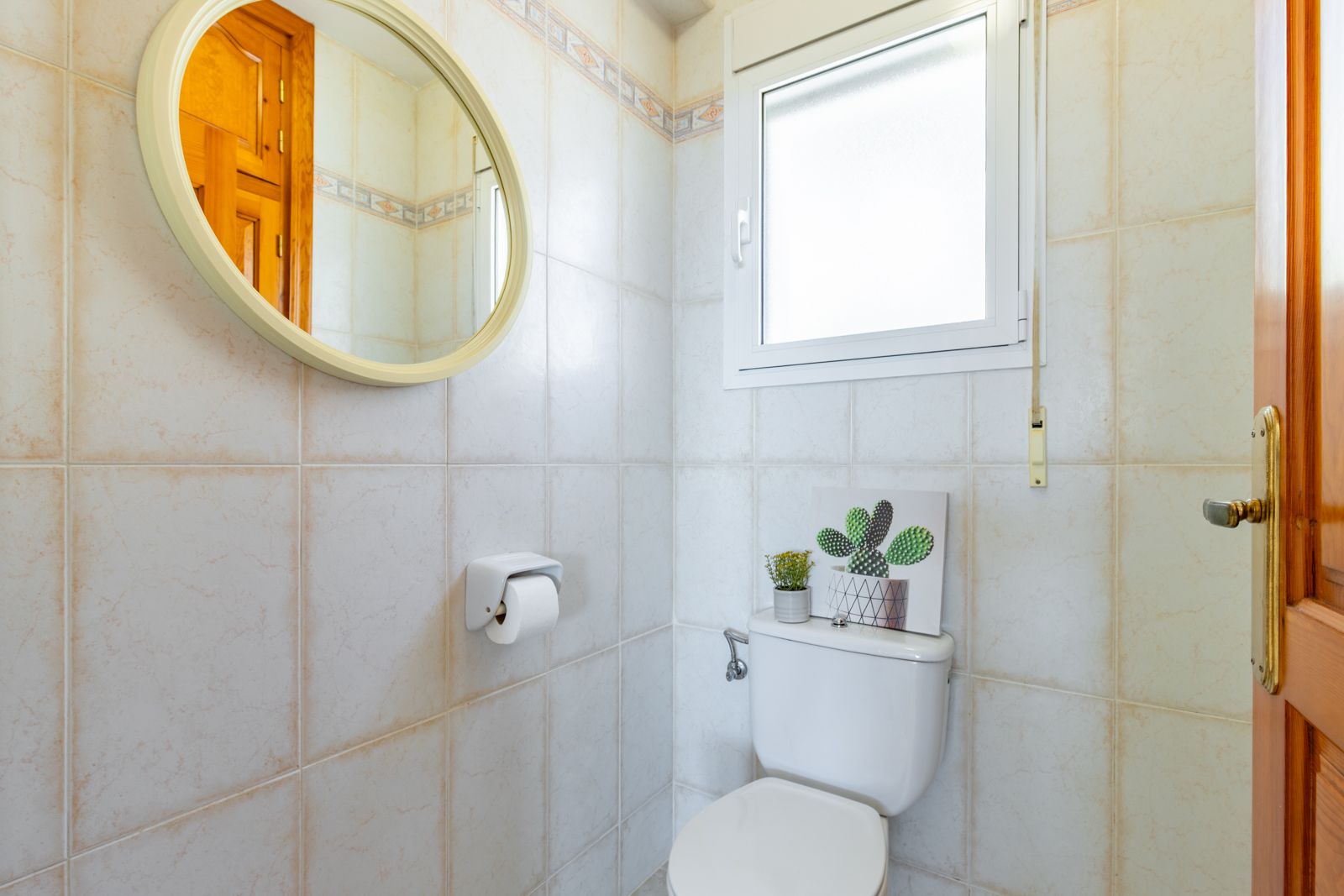 Functioneel gastentoilet met beige wandtegels, een ronde spiegel met licht frame en een raam met matglas voor natuurlijk licht en ventilatie.