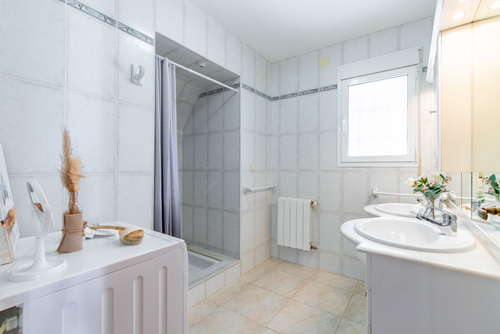 Functionele badkamer met dubbele marmeren wastafel, wasruimte, inloopdouche en radiator, afgewerkt met klassieke witte tegels.