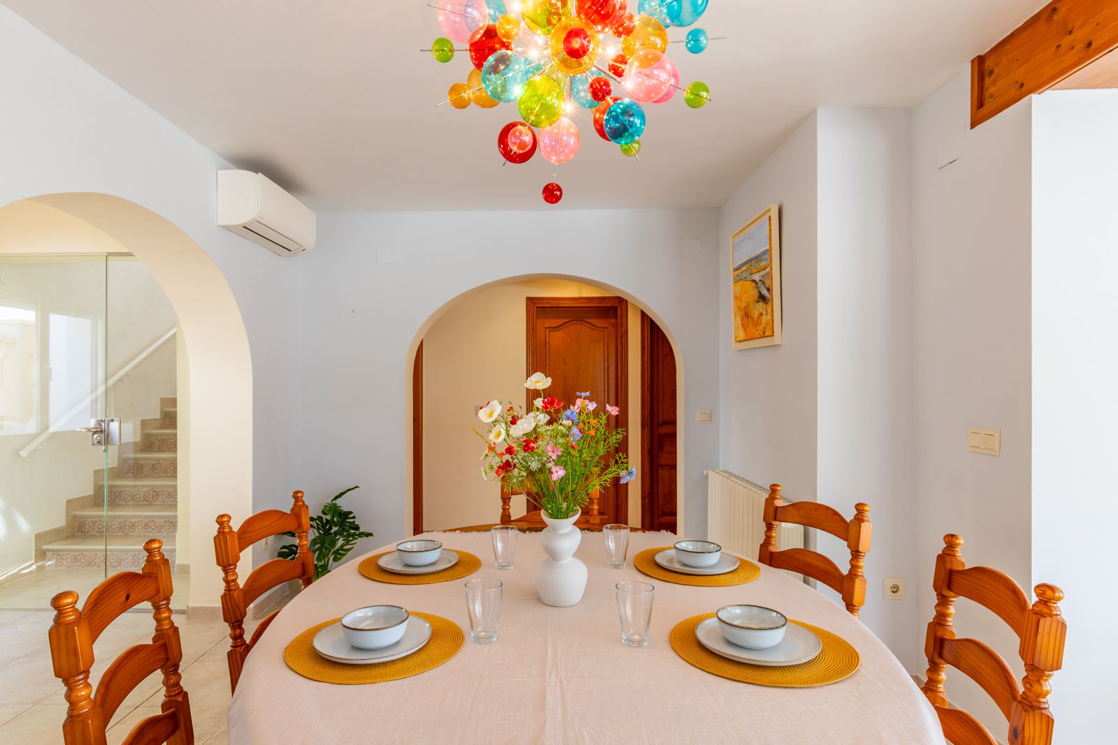 Lichte eetkamer met een ovale tafel, houten stoelen en een opvallende kleurrijke glazen bollenkroonluchter. De ruimte is voorzien van karakteristieke bogen en een betegelde trap.